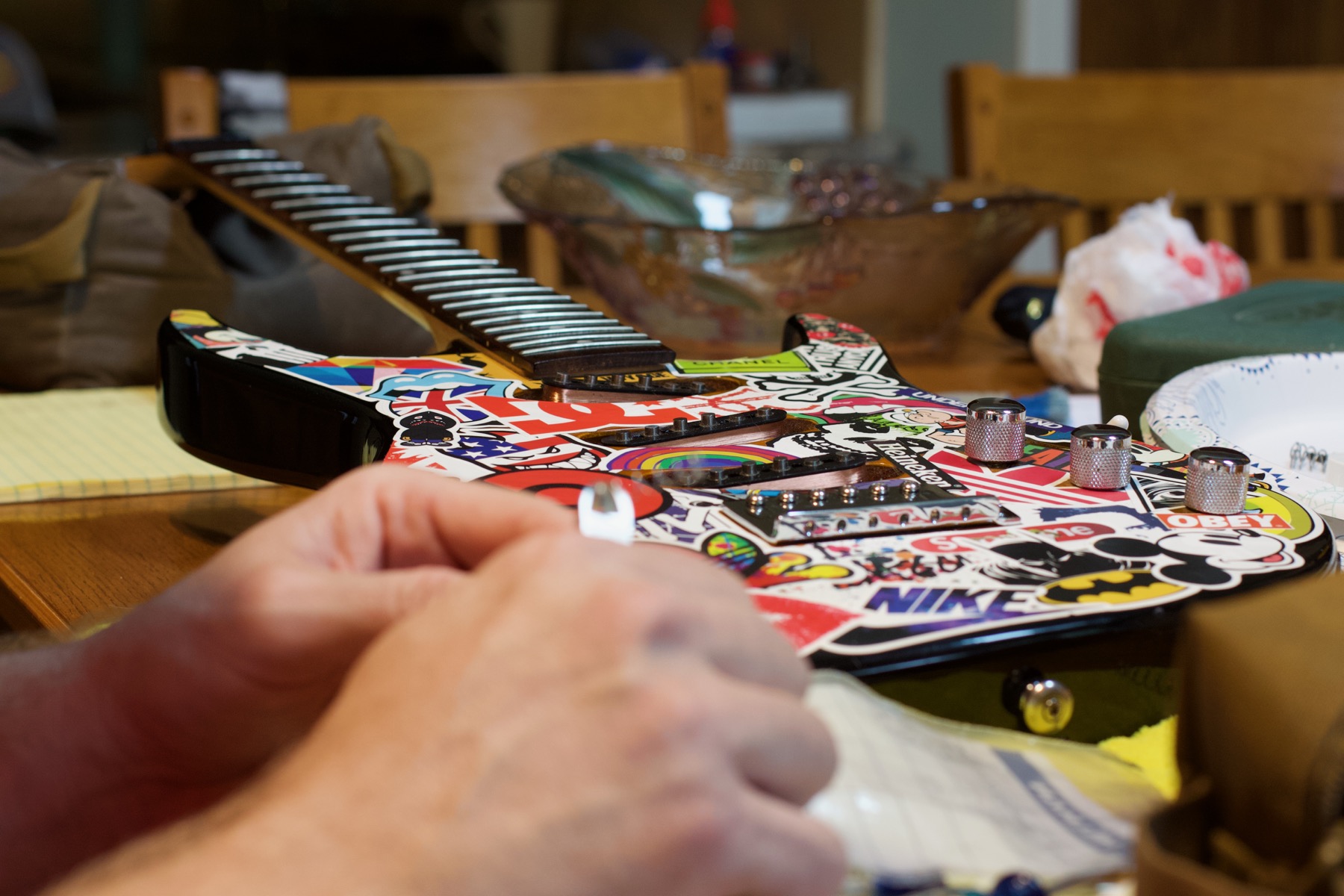 Hands working with small guitar parts on a kitchen table used as a teaching work surface.
